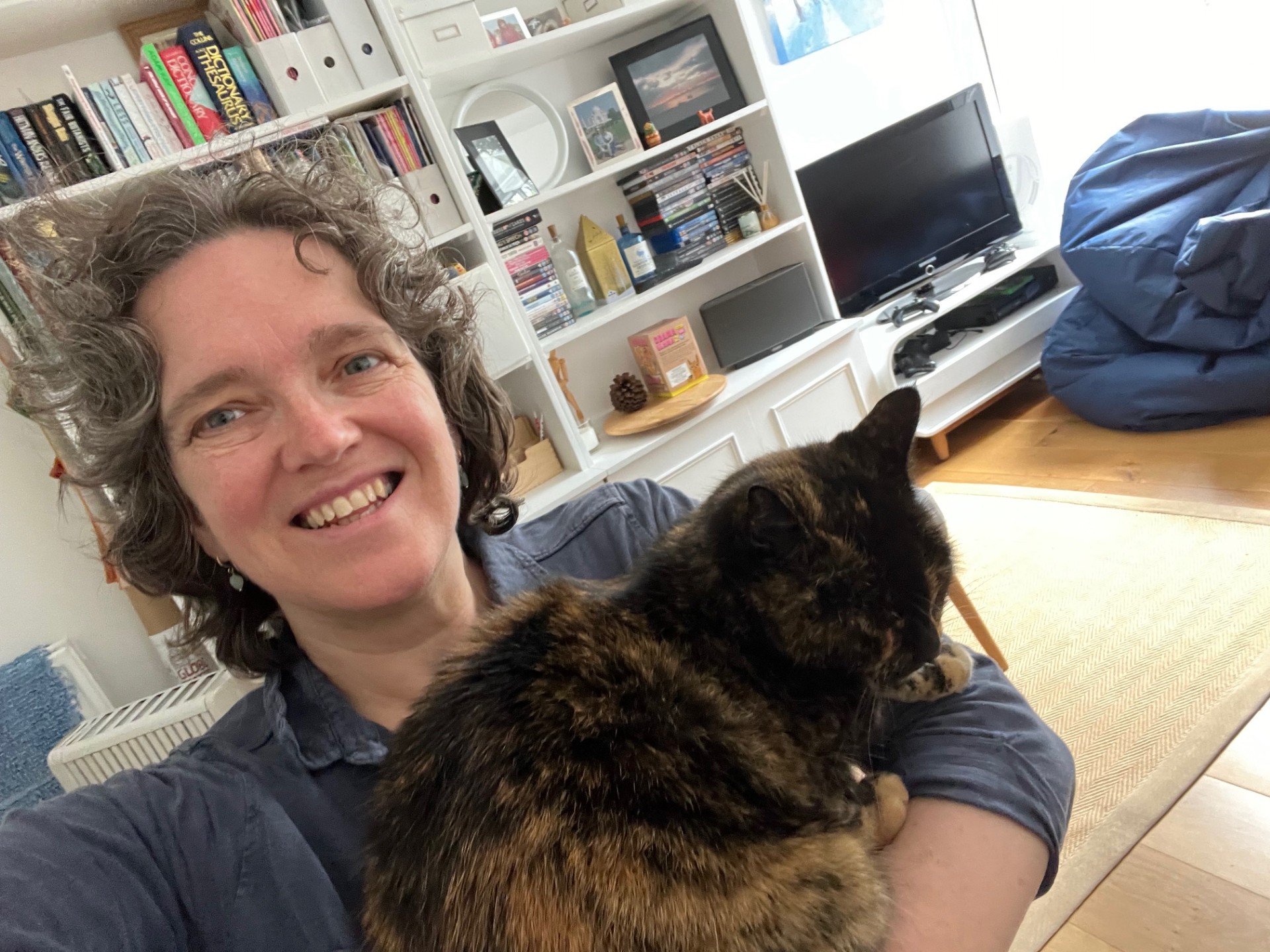 A person with curly hair smiles while holding a tortoiseshell cat. Behind them is a living room with a bookshelf, a television, and various items on shelves.